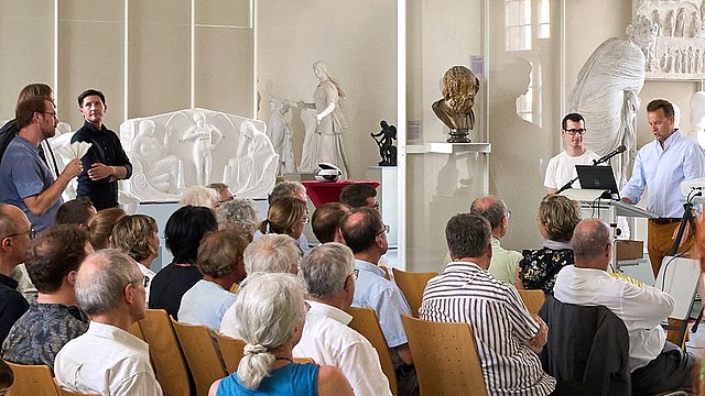 Blick schräg auf Stefan Krmnicek am Rednerpult mit dem sitzenden Publikum in der schrägen Rückansicht, die Stellwände des Rittersaals im Hintergrund.