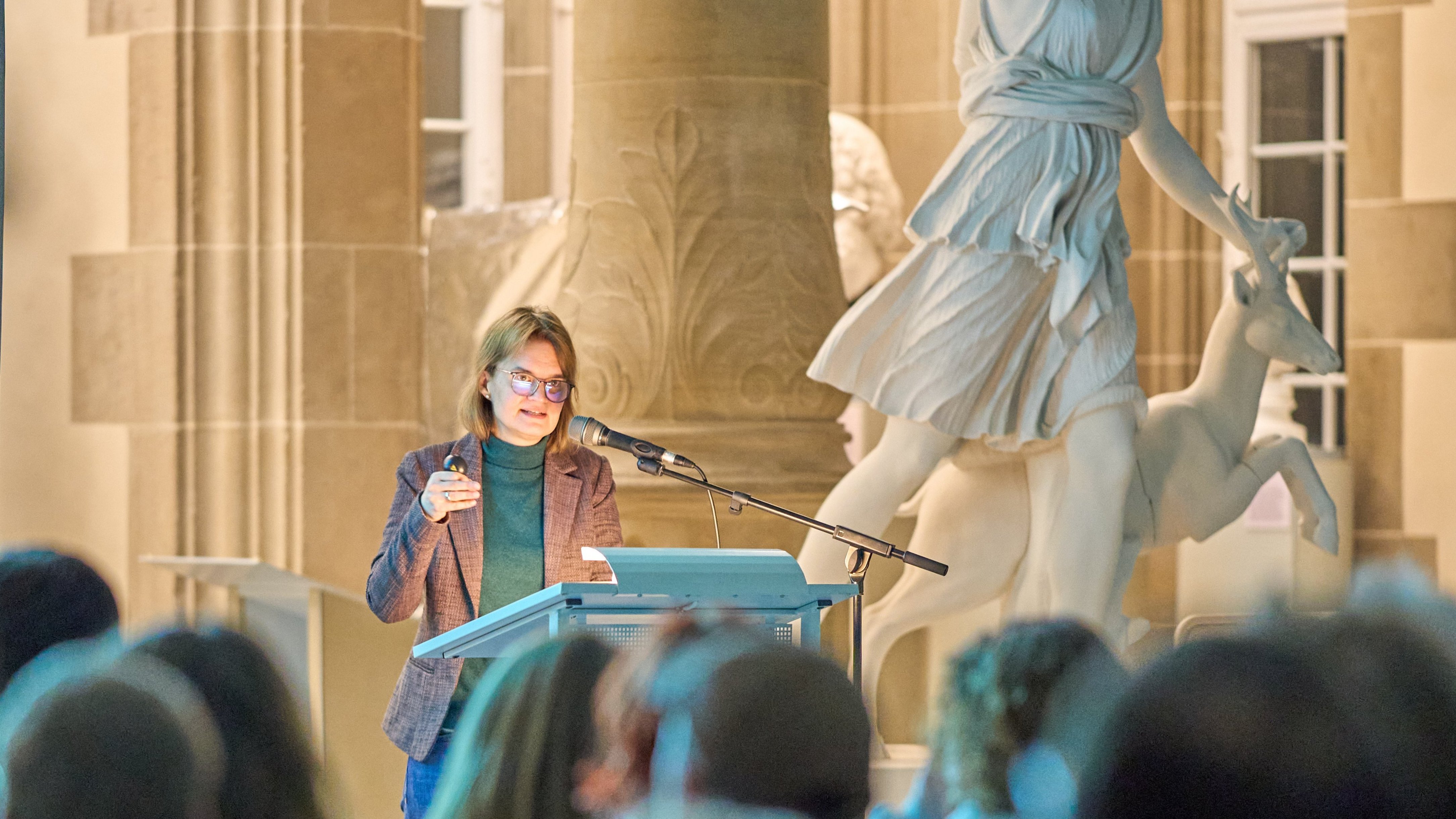 Dr Stephanie Walda-Mandel am Rednerpult der Eröffnung „Koloniale Schatten“ im Rittersaal; im Vordergrund Publikum von hinten