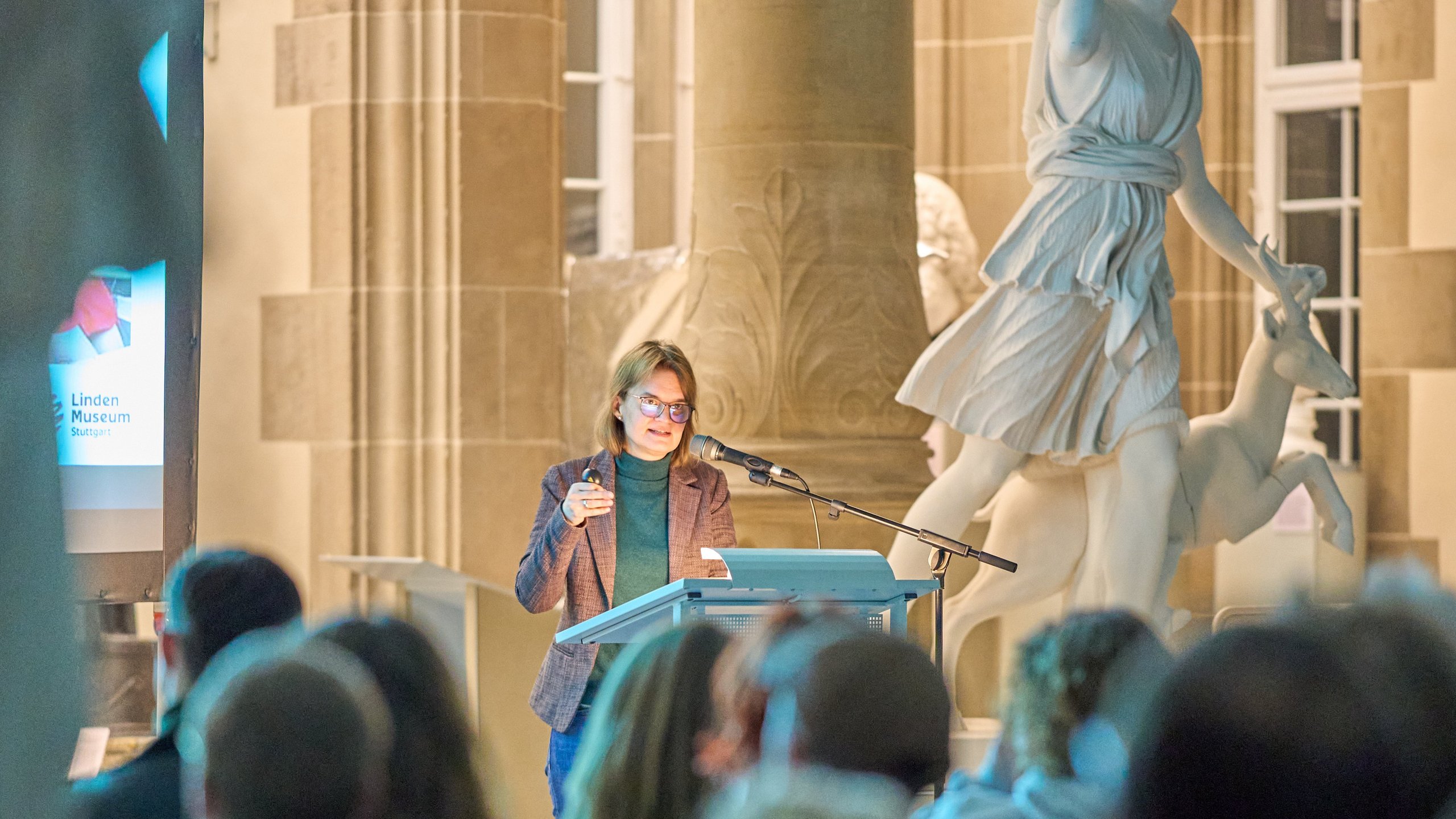 Dr Stephanie Walda-Mandel am Rednerpult der Eröffnung „Koloniale Schatten“ im Rittersaal; im Vordergrund Publikum von hinten