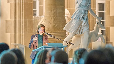 Foto: MUT | Fabian Kurze Dr Stephanie Walda-Mandel am Rednerpult der Eröffnung „Koloniale Schatten“ im Rittersaal; im Vordergrund Publikum von hinten