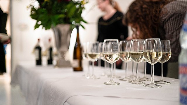 Flat view over a white-covered buffet with filled white wine glasses and a blurred background in which stands a goblet with a flower bouquet