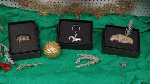 Foto: MUT | Nadja Mozdzen Auf grünem Tuch liegen drei schwarze Schächtelchen, umgeben von Zweigen falschen Immergrüns und einer goldenen Christbaumkugel; von links nach rechts sind in den Schächtelchen: eine elfenbeinfarbene Mammutreplik, ein silbernes Urzeitpferd als Schlüsselanhänger und eine elfenbeinfarbene Löwenreplik