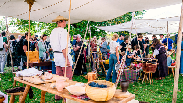 In den Zeltaufbauten drängt sich eine Menge aus Besuchenden, links im Bild ist ein Mann im weißen Gewand von hinten zu sehen, der hinter einer Bank mit Alltagsgegenständen wie einer Schüssel mit Beeren steht