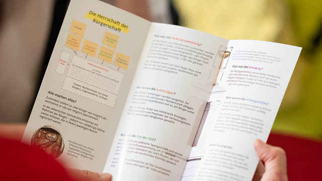 Blick über die Schulter einer Person in roter Kleidung, die in den Händen einen Flyer der Ausstellung "Agora" aufgeschlagen hat: einen dreiseitigen Folder mit einem Schaubild der athenischen Verfassung und Detailtexten zu verschiedenen Institutionen sowie Fotos einer Silbermünze und eines tempelartigen Denkmals