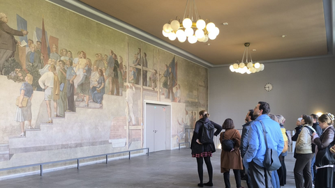 Foto: MUT| Michael La Corte Rechts ist eine Gruppe von Menschen, die sich schauend gen links neigen, wo auf einer Wand freskenartig eine Tribüne mit Zuschauenden gemalt ist