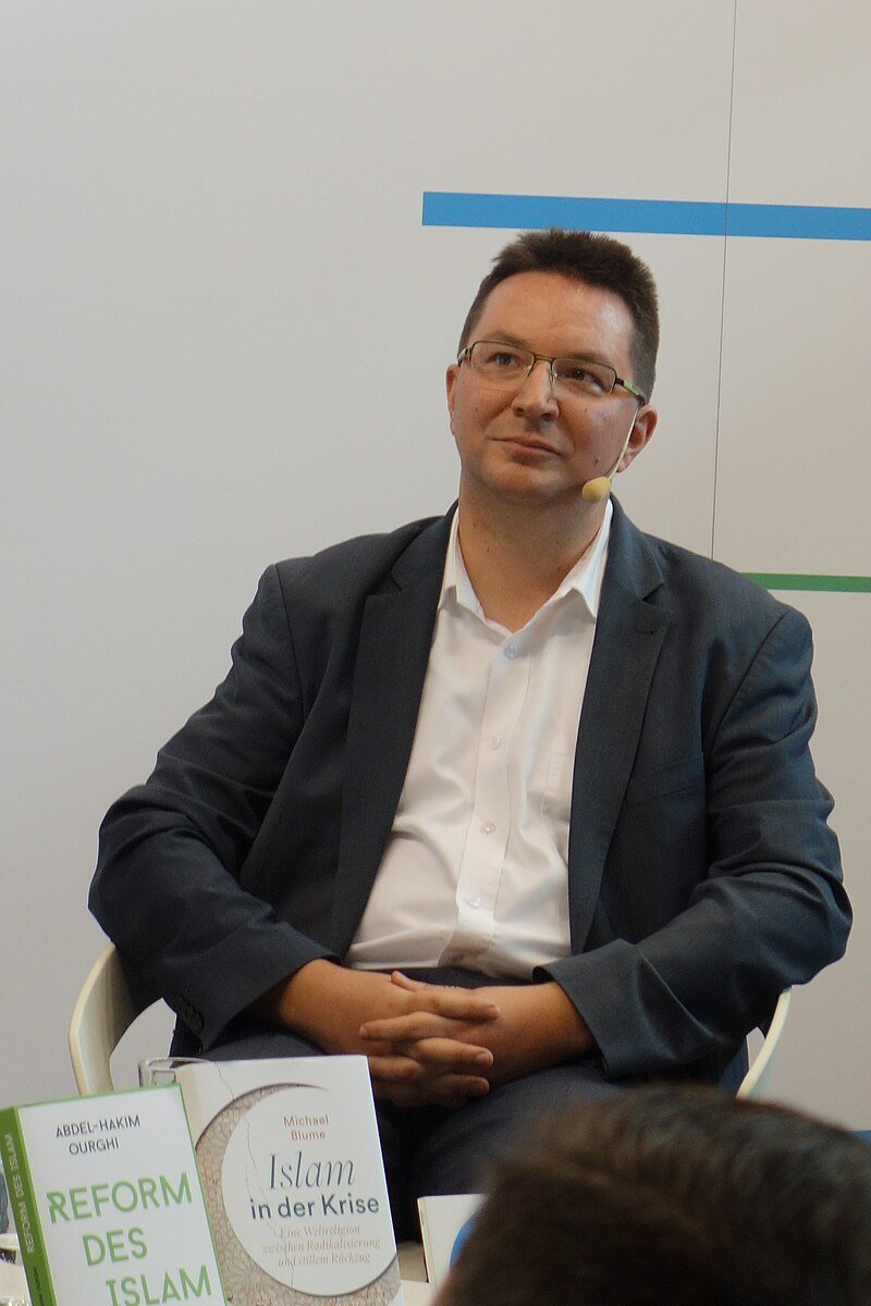 Ein Mann mittleren Alters sitzt in einem weißen Stuhl vor einer hellen Wand mit horizontalen blauen und grünen Linien. Er trägt eine Brille, ein weißes Hemd und einen dunklen Anzug. Er hat ein Headset mit Mikrofon am rechten Ohr. Seine Hände sind locker vor ihm verschränkt. Vor ihm stehen zwei Bücher mit den Titeln „REFORM DES ISLAM“ und „Islam in der Krise“. Im Vordergrund ist unscharf der Kopf einer Person zu sehen.