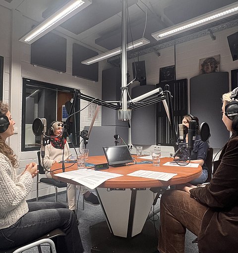 Aufnahme im Tonstudio: Vier Personen sitzen um einen runden Tisch mit Mikrofonen, die den Podcast aufnehmen.