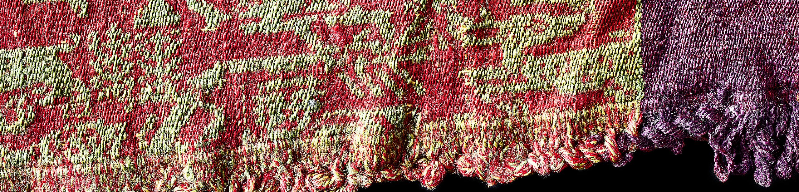 Close-up of a late antique textile with a visible selvedge against a black background; on the left, red with a green animal pattern; on the right, purple