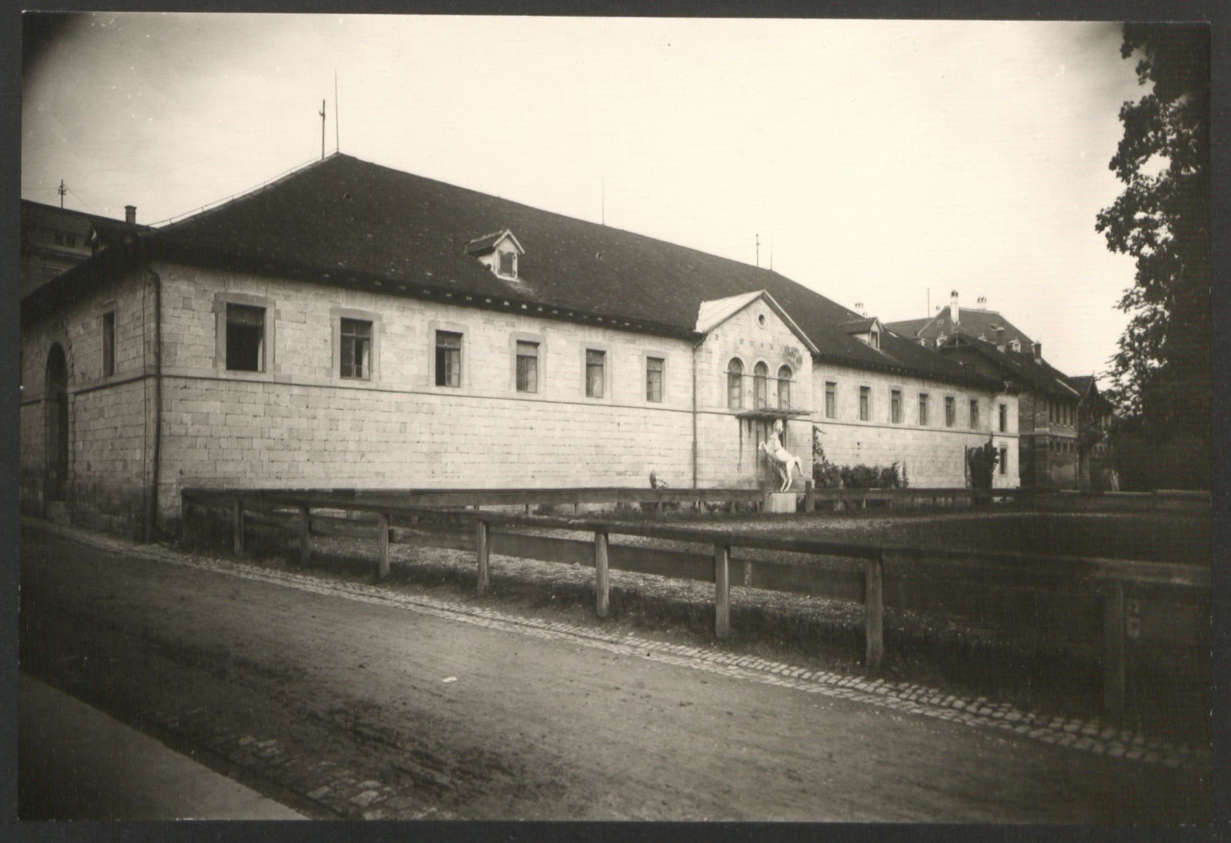 Reitstall, 1905 Vignettiertes Schwarz-weiß-Foto eines langgestreckten hellen Baus mit dunklem Dach und der Statue eines sich aufbäumenden Pferdes vor dem zentralen Eingang