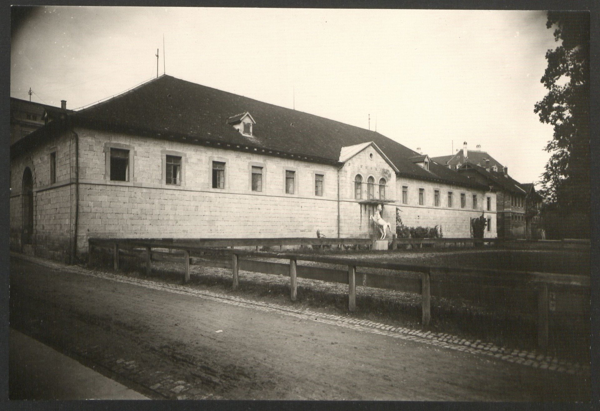 Reitstall, 1905 Vignettiertes Schwarz-weiß-Foto eines langgestreckten hellen Baus mit dunklem Dach und der Statue eines sich aufbäumenden Pferdes vor dem zentralen Eingang