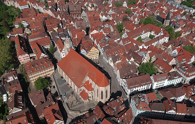 Luftaufnahme einer historischen Altstadt mit roten Ziegeldächern und engen Gassen. Im Zentrum befindet sich ein großes rechteckiges Gebäude mit einem Innenhof, das von einem hohen Turm mit spitzem Dach überragt wird. Umgeben ist das Areal von zahlreichen anderen Gebäuden und Straßen. Die Architektur wirkt mittelalterlich und gut erhalten.