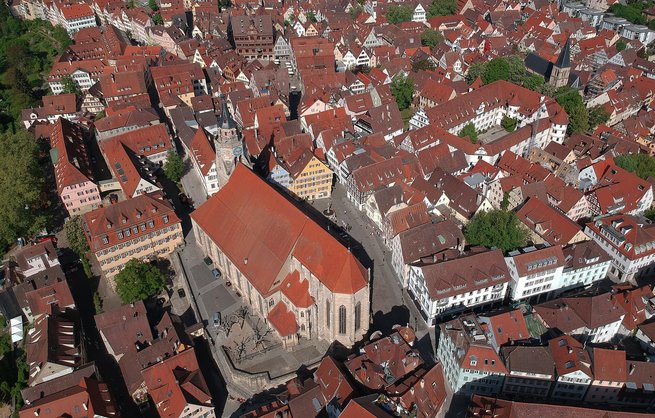 Luftaufnahme einer historischen Altstadt mit roten Ziegeldächern und engen Gassen. Im Zentrum befindet sich ein großes rechteckiges Gebäude mit einem Innenhof, das von einem hohen Turm mit spitzem Dach überragt wird. Umgeben ist das Areal von zahlreichen anderen Gebäuden und Straßen. Die Architektur wirkt mittelalterlich und gut erhalten.