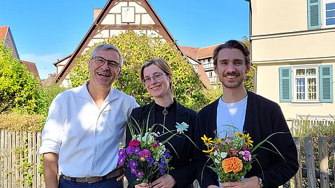 Ernst Seidl (links), Nadja Mozdzen (mittig) und Simon Zauner (rechts), letztere beide mit Blumensträußen in der Hand, stehen bei sonnigem Wetter vor der Kulisse der Tübinger Altstadt