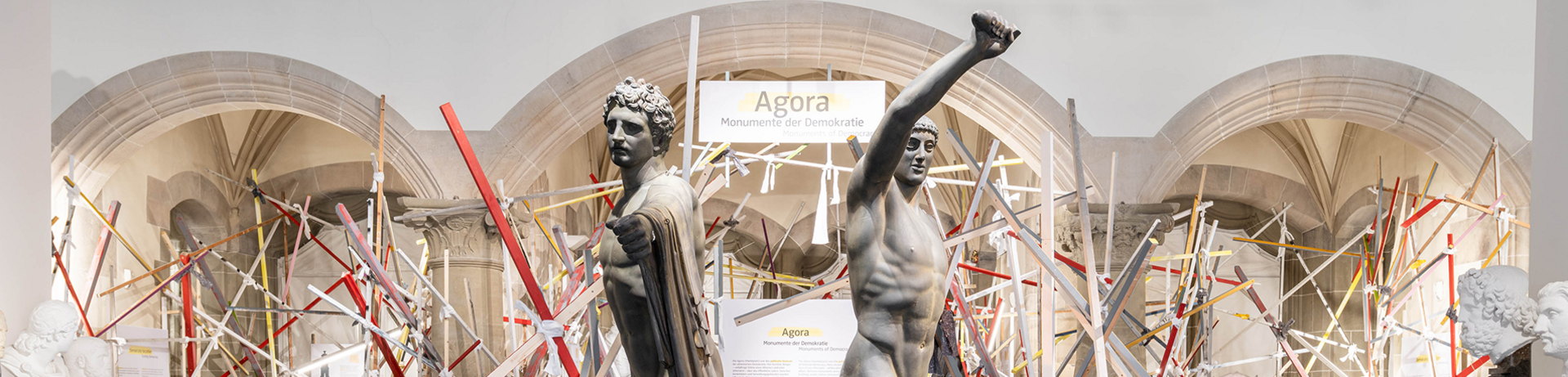 wide panoramic view of the special exhibition ‘Agora’ in the niches of the Knights’ Hall, featuring a multicoloured framework of wooden beams of varying sizes, with the casts of the Tyrannicides, arms raised in a fighting stance, shown head-on in the foreground 