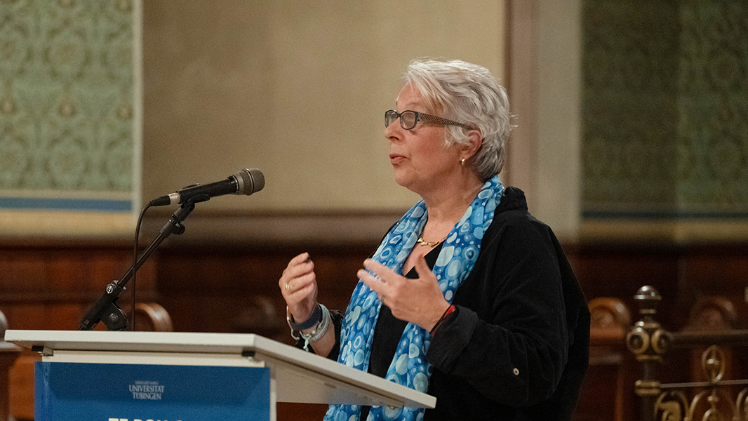 Am Rednerpult mit dem blauen Ausstellungsplakat von "Te Pou o Hinematioro" steht gestikulierend Professorin Gabriele Alex