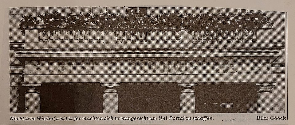 Schwarz-weißes Foto eines historischen Gebäudeportals. Über dem Eingang ist in großer, gesprühter Schrift „ERNST BLOCH UNIVERSITÄT“ zu lesen. Die Buchstaben sind ungleichmäßig und wirken wie ein nächtliches Graffiti. Über dem Graffito stehen Blumenkästen auf dem Balkongeländer.  Am unteren Bildrand befindet sich ein erklärender Text: „Nächtliche Wiedertäufer machten sich termingerecht am Uni-Portal zu schaffen.“ Rechts daneben: „Bild: Gööck“
