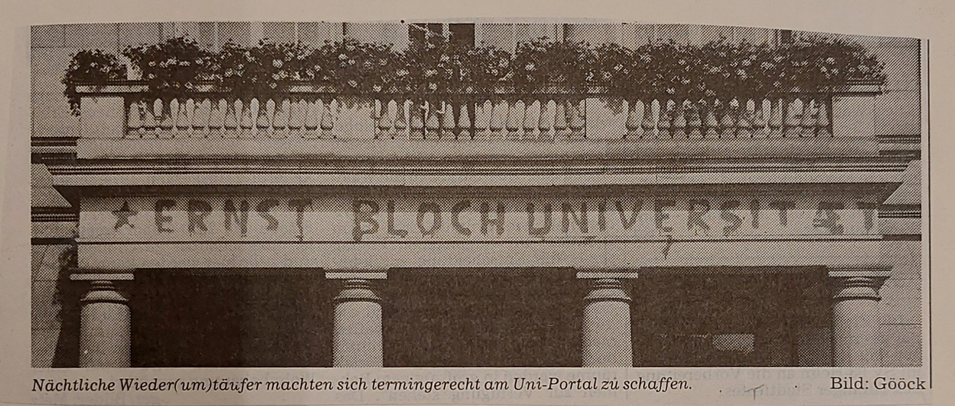 Schwarz-weißes Foto eines historischen Gebäudeportals. Über dem Eingang ist in großer, gesprühter Schrift „ERNST BLOCH UNIVERSITÄT“ zu lesen. Die Buchstaben sind ungleichmäßig und wirken wie ein nächtliches Graffiti. Über dem Graffito stehen Blumenkästen auf dem Balkongeländer. Am unteren Bildrand befindet sich ein erklärender Text: „Nächtliche Wiedertäufer machten sich termingerecht am Uni-Portal zu schaffen.“ Rechts daneben: „Bild: Gööck“