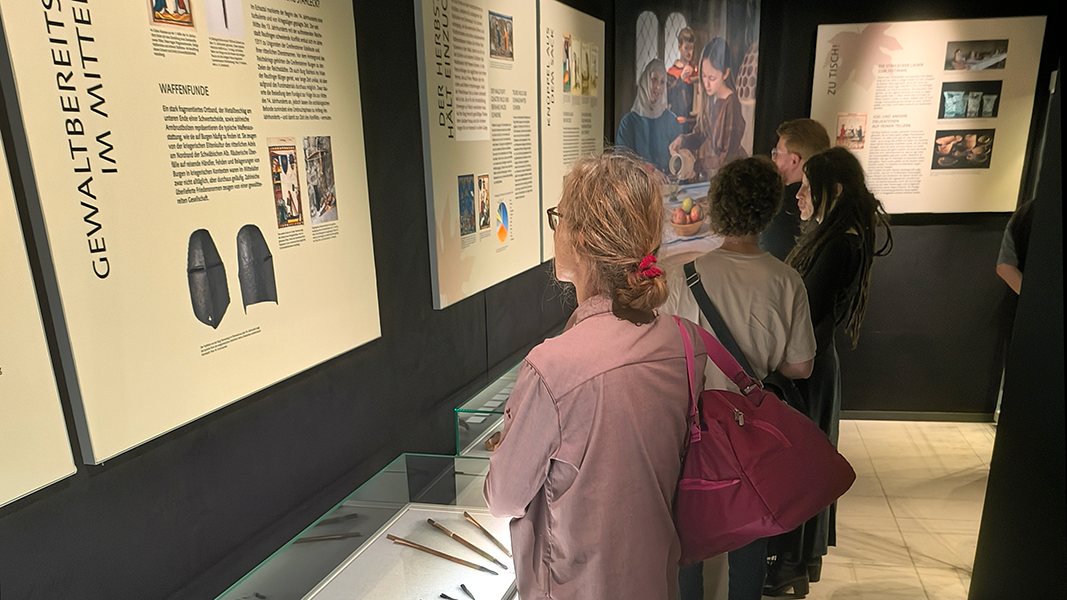 Besuchende stehen in der Ausstellung vor einer Reihe an beleuchteten Tischvitrinen und sehen auf die Tafeln darüber an der Wand; im Hintergrund ist ein großes Bild mit mittelalterliche gekleideten Leuten zu sehen