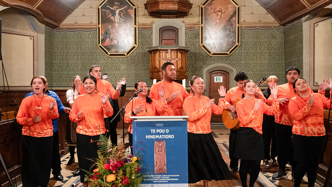 Hinter dem Rednerpult mit dem blauen Ausstellungsplakat von "Te Pou o Hinematioro" tanzen in zwei Reihen die Schüler*innen der Te Aitanga-a-Hauiti in zwei Reihen aufgestellt; alle haben mit angewinkelten Armen die Hände zu ihrer Linken erhoben