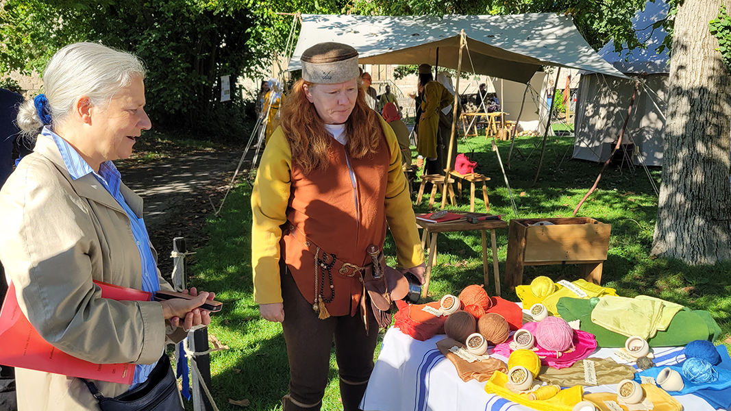 Universitätsrektorin Professorin Dr. Dr. h.c. (Doshisha) Karla Pollmann, links im Bild, betrachtet einen Tisch mit gefärbten Wollbeispielen; neben ihr eine Frau in orangenem, gegürtetem Wams mit gelben Ärmeln und dunklen Hosen