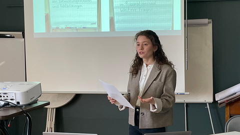 Foto: MUT | Annika Vosseler Dielleza Hyseni gestikulierend im Vortrag vor einem Ausschnitt der Präsentationsfolie mit Fotos von beschrifteten Stellwänden