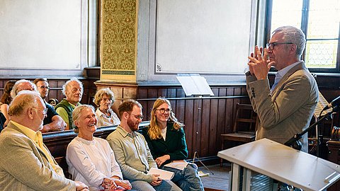 Professor Dr. Ernst Seidl, rechts, gestikuliert nach links gewandt, wo in der ersten Reihe der Schlosskirche unter anderem Rektorin Professorin Dr. Dr. hc. Karla Pollmann, Lucas Rau und Teresa Pohl sitzen (vlnr)