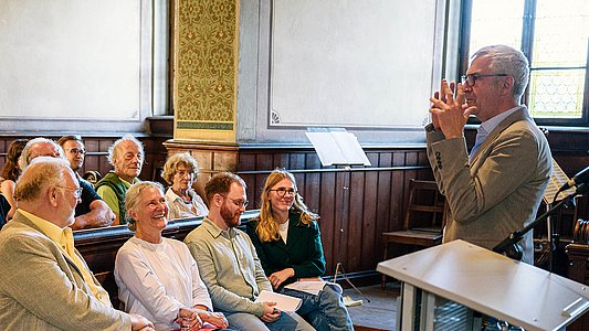 Professor Dr. Ernst Seidl, rechts, gestikuliert nach links gewandt, wo in der ersten Reihe der Schlosskirche unter anderem Rektorin Professorin Dr. Dr. hc. Karla Pollmann, Lucas Rau und Teresa Pohl sitzen (vlnr)