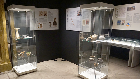 Foto: MUT | Michael La Corte Blick in die Ausstellung "Ausgegraben!" im Kabinettraum des MUT | Museum Alte Kulturen mit schwarz verkleideten Wänden und hellen Wandtafeln, davor zwei beleuchtete Standvitrinen aus Glas mit mehreren Ebenen, auf denen Keramikfunde liegen; rechts im Hintergrund sind beleuchtete Tischvitrinen zu erkennen