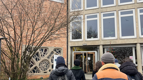 Am unteren Bildrand in Rückansicht die Studierenden des Seminars, die auf ein Gebäude mit Betonfassade (rechts) und Backstein (links) zugehen. In der Backsteinfassade ist im Erdgeschoss das Buntglasfenster in organischen Formen sichtbar, das zum Andachtsraum der Klinik gehört