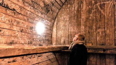 Eine Person steht im Inneren des Fasses und richtet eine Taschenlampe auf die bekritzelte Innenwand. Der weiße Lichtkegel erscheint auf dem Holz