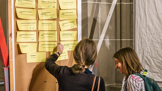 Zwei Besucherinnen stehen vor einer Pinnwand links im Bild, eine zeigt auf einen der gelben Moderationszettel, auf denen Charaktereigenschaften stehen, die einem guten Redner in der Antike zugeschrieben wurden; grüne Punkte markieren vereinzelte Zettel