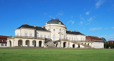 Fotografie eines symmetrisch aufgebauten Barockschlosses mit heller Fassade, zentraler Kuppel und ausladenden Seitenflügeln. Vor dem Schloss erstreckt sich eine große Rasenfläche. Der Himmel ist klar und blau mit wenigen kleinen Wolken. Die Architektur wirkt monumental und repräsentativ.