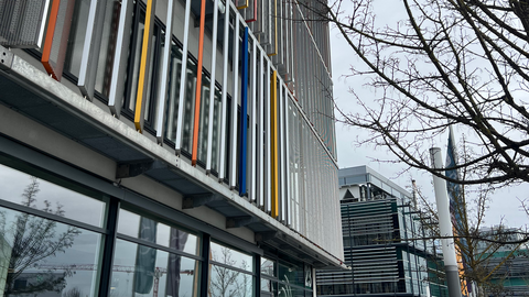 Blick auf eine Fassade aus leichter Froschperspektive, die über das verglaste Erdgeschoss hervorsteht und mit farbig gefassten, schmalen Rechtecken aus Metallgitter verdeckt wird