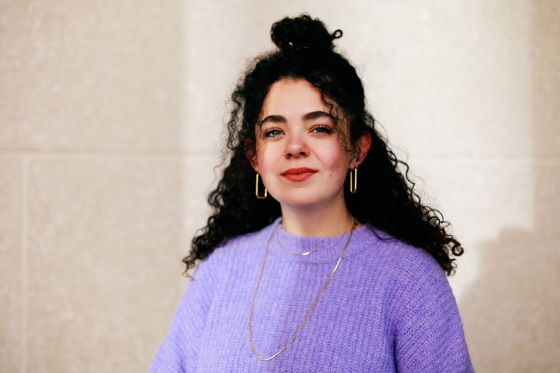 Eine junge Frau mit lockigem, dunklem Haar, das teilweise zu einem Dutt gebunden ist, steht vor einer hellen Wand. Sie trägt einen fliederfarbenen Strickpullover, goldene rechteckige Ohrringe und zwei zarte Goldketten. Ihr Gesicht ist leicht geschminkt, sie lächelt dezent und schaut direkt in die Kamera.