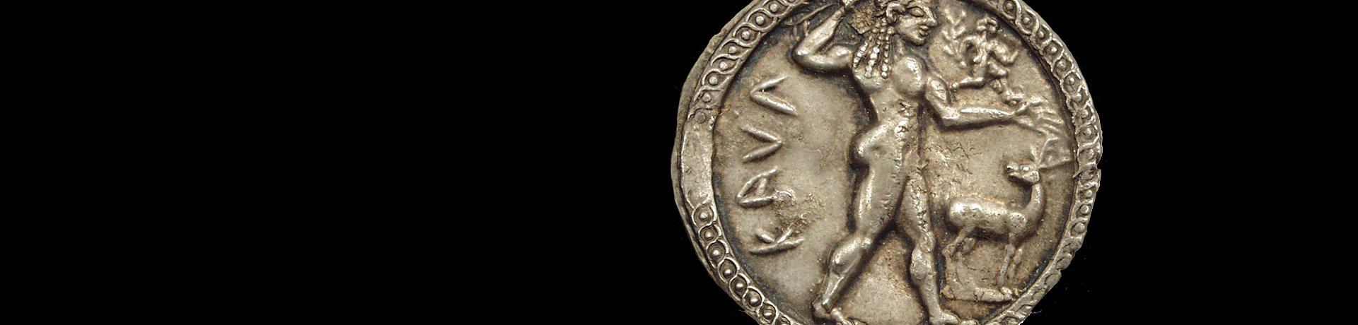 Foto: MUT Bildseite (Avers) einer Silbermünze mit einem jungen, nach rechts schreitenden Apollon, auf dessen ausgestreckten Arm eine kleine, männliche Figur nach rechts rennt; zur Rechten des Gottes wendet sich ein kleiner Hirsch nach ihm um; links des Gottes steht die griechische Inschrift "KAUL" (für Kaulonia)
