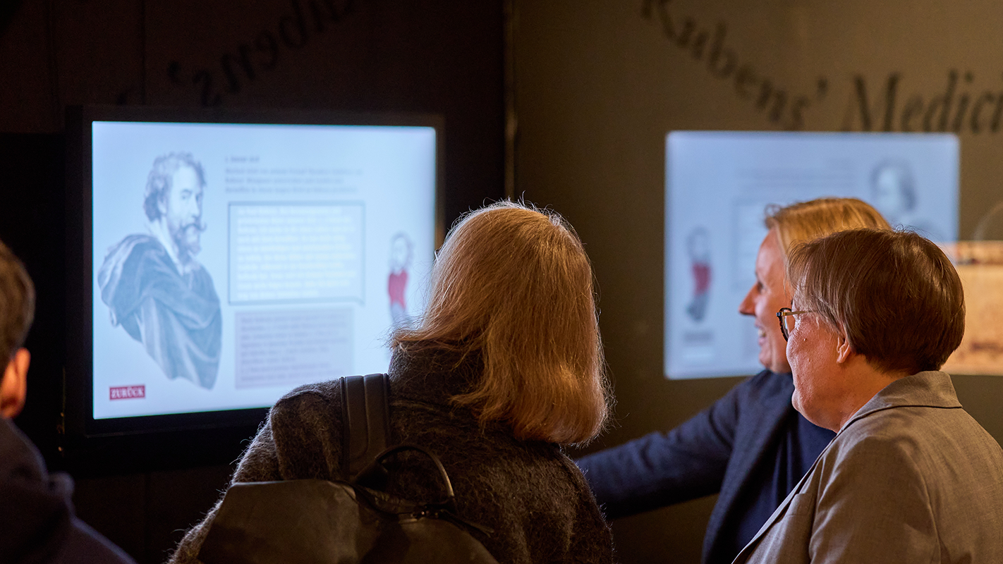 Besuchende in der Rückansicht vor einem an der Wand montierten Touchscreen, aber dem ein interaktiver Dialog angezeigt wird