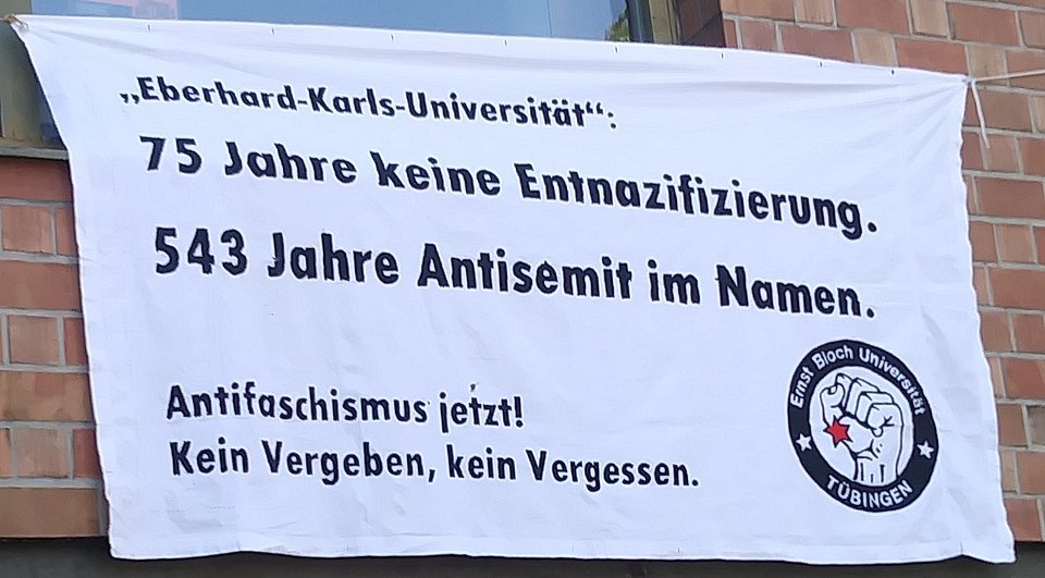 Ein weißes Protestbanner hängt an einer Backsteinwand. In schwarzer, fettgedruckter Schrift steht darauf:  „‚Eberhard-Karls-Universität‘: 75 Jahre keine Entnazifizierung. 543 Jahre Antisemit im Namen.  Antifaschismus jetzt! Kein Vergeben, kein Vergessen.“  Rechts unten befindet sich ein rundes Logo mit einer erhobenen Faust, die einen roten Stern in der Mitte zeigt. Im Kreis um das Bild steht: „Ernst Bloch Universität Tübingen“.