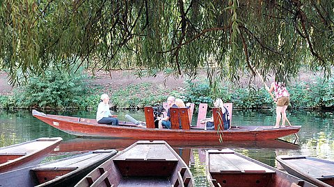 Blick vom Ufer auf den Neckar und einen zentral durch das Bild fahrenden Stocherkahn mit Frau Rektorin Prof. Dr. Karla Pollmann (links), die in eine Kamera spricht, oben gerahmt von Weidenästen, unten von anlegenden Stocherkähnen.