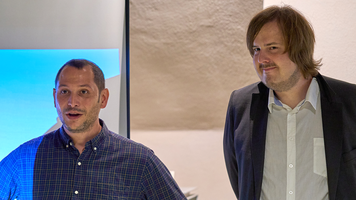 Links ist Dr. Michael La Corte im dunklen, karierten Hemd vor der blauen Projektion einer Präsentation zu sehen, rechts Kevin Körner in weißem Hemd und schwarzer Anzugjacke