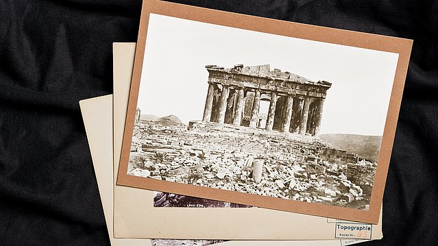 Auf einem Stapel von drei alten Fotographien liegt ein Foto der Ruine eines Rundtempels obenauf