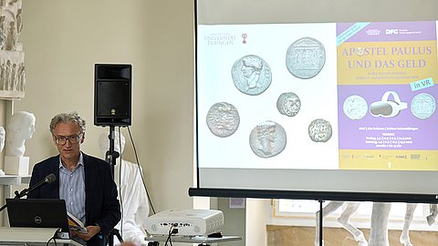 Links Jörg Robert am Rednerpult, hinter ihm eine Leinwand mit Titel und Plakat der VR-Ausstellung "Apostel Paulus und das Geld"