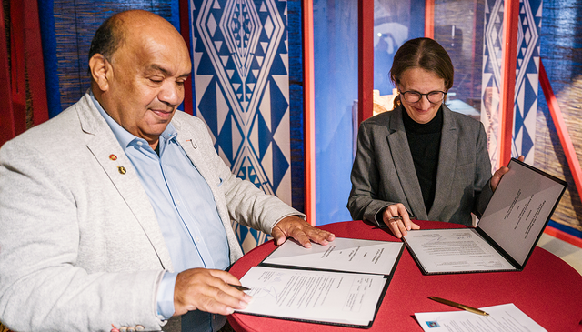 Victor Walker (links) und Monique Scheer (rechts) unterzeichnet an einem Stehtisch mit roter Husse die Rückgabevereinbarung