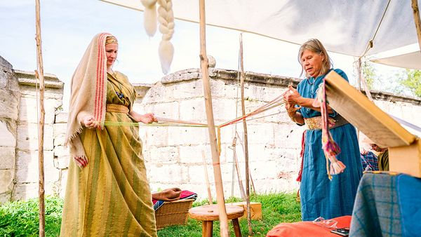 Zwei Frauen in langen, gelben beziehungsweiser blauen Gewändern flechten gemeinsam an einem einfachen Holzgestell Band