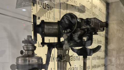 Hell erleuchtetes historisches Gerät in einer spiegelnden Glasvitrine mit dem Oberteil eines Metallgefäßes links und einem röhrenartigen Sehapparat rechts