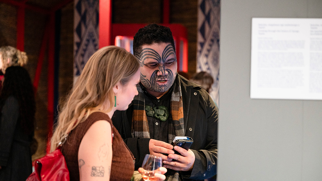 Vor einer Vitrine, deren Inhalt nicht zu erkennen ist, stehen eine Besucherin und ein Gast der Delegation der Te Aitanga-a-Hauiti mit Gesichtstattoos