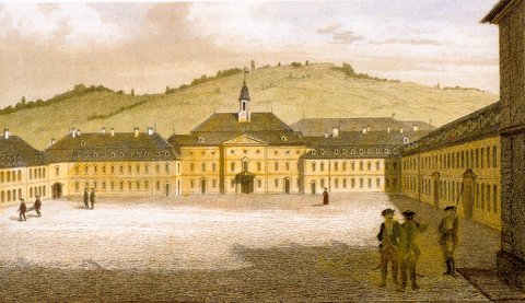 Historische Darstellung eines weitläufigen Platzes mit zentralem Gebäude im Barockstil, das einen Uhrturm trägt. Mehrere Personen in historischer Kleidung gehen oder stehen auf dem Platz. Im Hintergrund erstrecken sich sanfte, grüne Hügel. Rechts im Bildrand ist ein weiteres Gebäude zu sehen. Die Szene wirkt ruhig und idyllisch, möglicherweise handelt es sich um eine Darstellung aus dem 18. oder frühen 19. Jahrhundert.