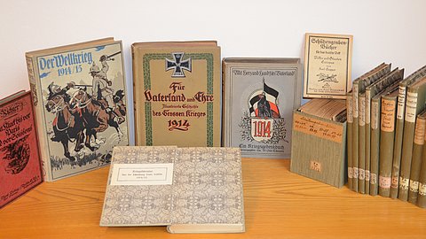 Foto: Universitätsbibliothek Tübingen Auf einem Holztisch aufgestellte Bände der Kriegsliteratur Laiblin mit mehrheitlich olivgrünem Buchbindereinband; vor dem im Halbrund aufgestellten Büchern zeigt ein aufgeschlagenes Buch seinen gemusterten Vorsatz