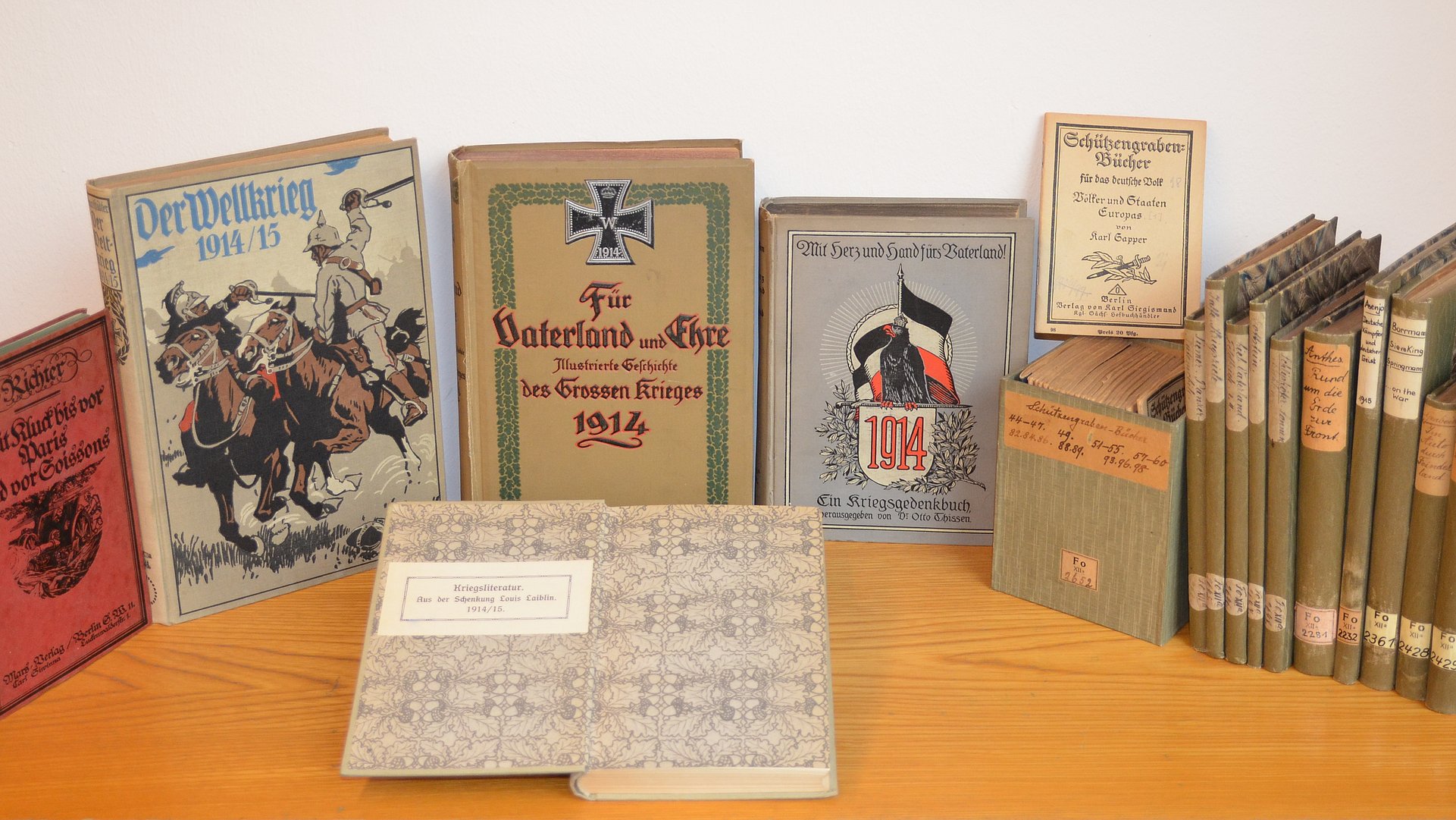 Foto: Universitätsbibliothek Tübingen Auf einem Holztisch aufgestellte Bände der Kriegsliteratur Laiblin mit mehrheitlich olivgrünem Buchbindereinband; vor dem im Halbrund aufgestellten Büchern zeigt ein aufgeschlagenes Buch seinen gemusterten Vorsatz