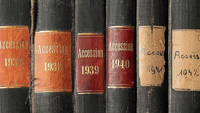 Detailaufnahme einer Reihe dunkler Buchrücken mit zunächst gold auf braun geprägten, in den letzten drei Bänden handbeschrifteten  Schildern mit "Accession" und den fortlaufenden Jahreszahlen von 1936 bis 1943. Die Schilder der jüngeren Bände sind deutlich einfacher und abgenutzter.