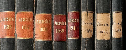 Detailaufnahme einer Reihe dunkler Buchrücken mit zunächst gold auf braun geprägten, in den letzten drei Bänden handbeschrifteten  Schildern mit "Accession" und den fortlaufenden Jahreszahlen von 1936 bis 1943. Die Schilder der jüngeren Bände sind deutlich einfacher und abgenutzter.